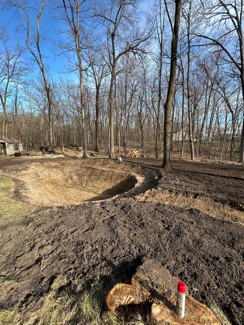Excavated area in a forested yard, surrounded by bare trees and dirt piles.