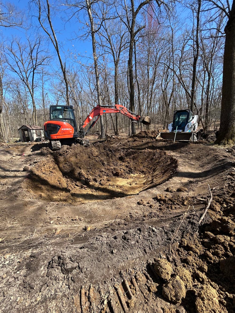 Excavation site with heavy machinery digging in a forested area, showcasing a large dirt pit.