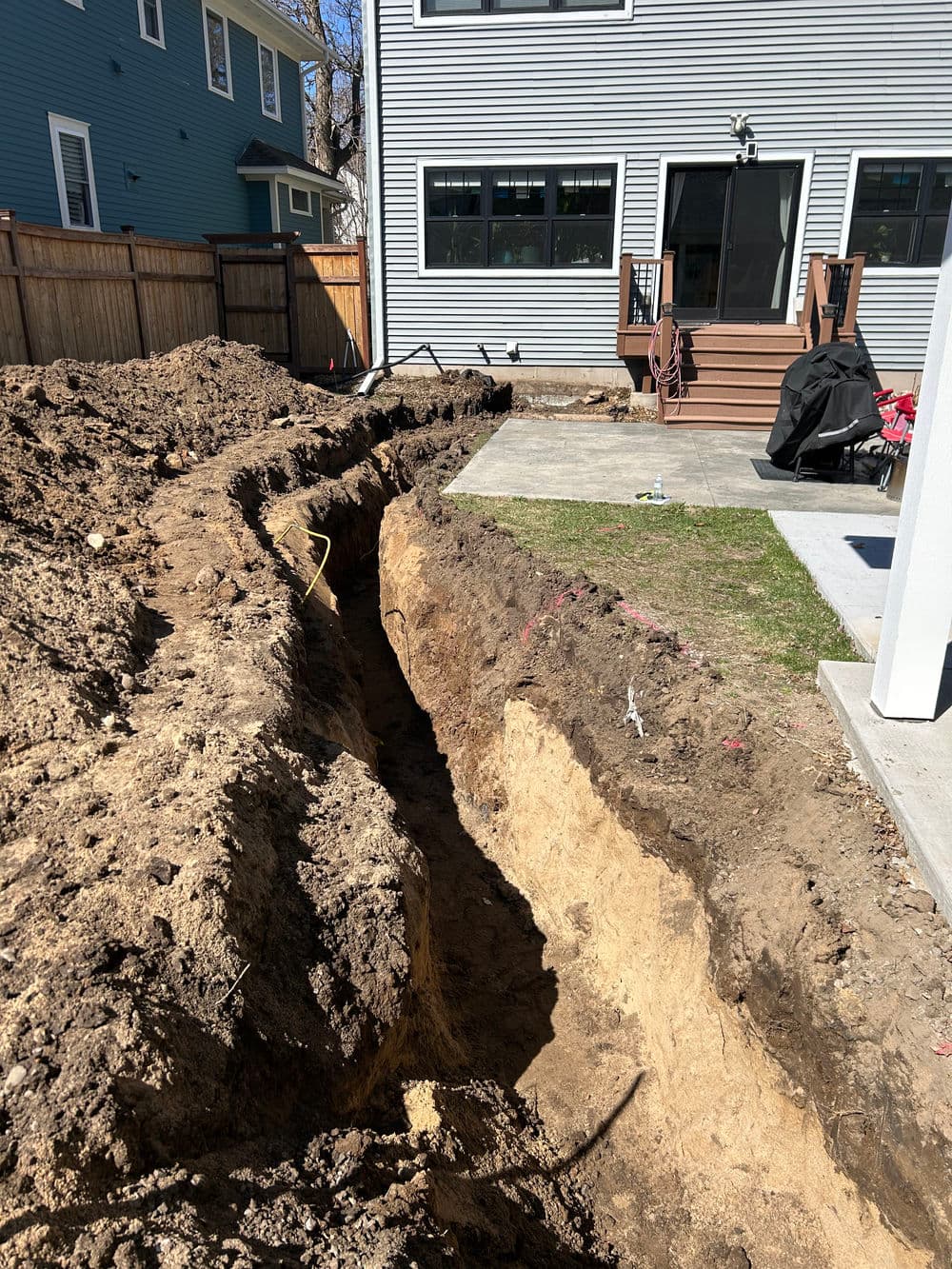 Excavated trench in backyard for construction near house with deck and concrete patio.