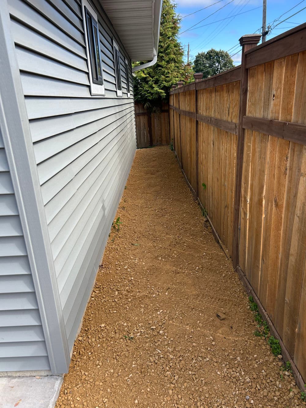 Narrow gravel pathway between a house and wooden fence in a residential area.