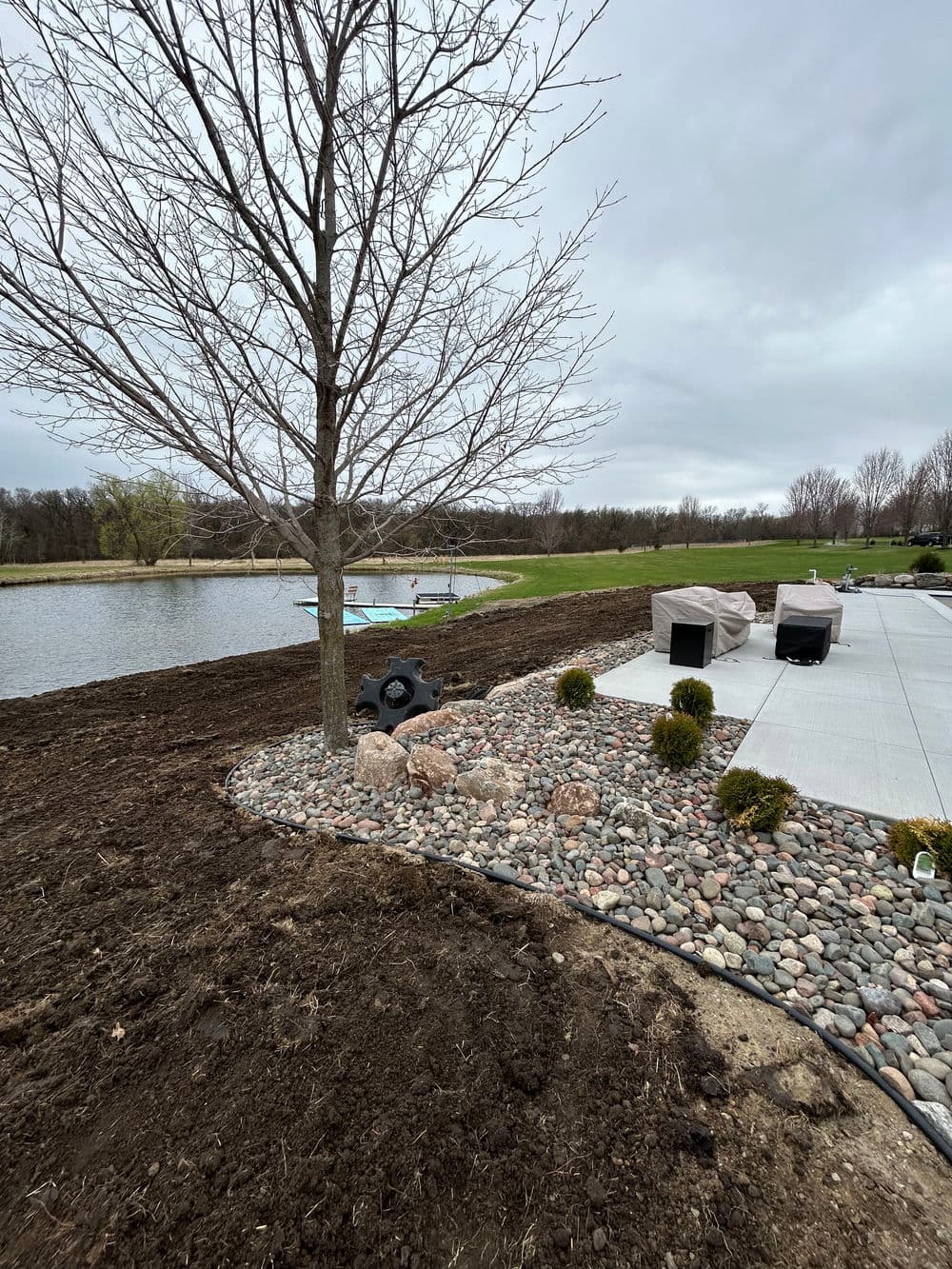 Lakeside landscape with a tree, stones, and outdoor seating area by the water.