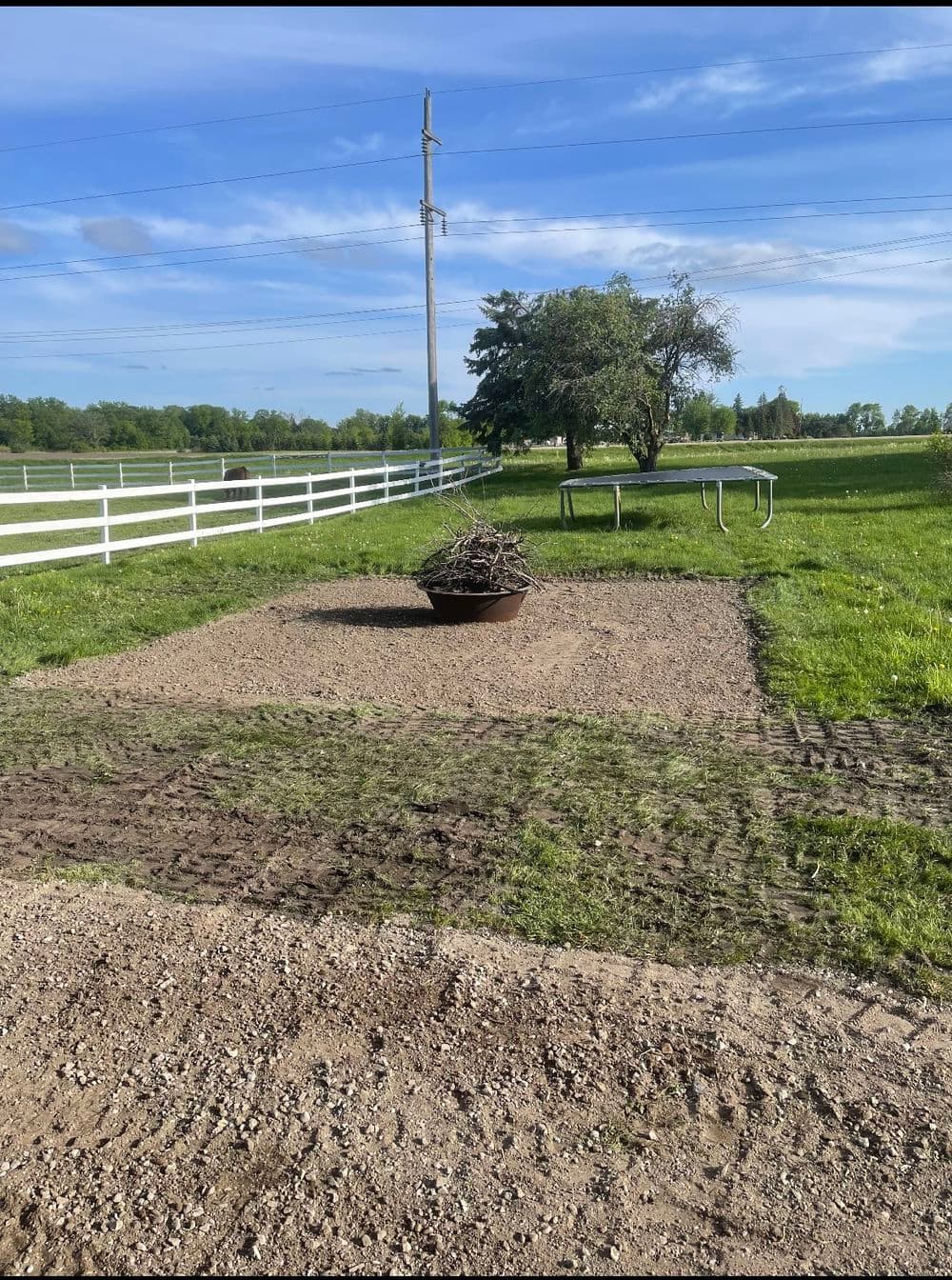 Field preparation with a pile of branches and gravel on a sunny day, white wooden fence in background.
