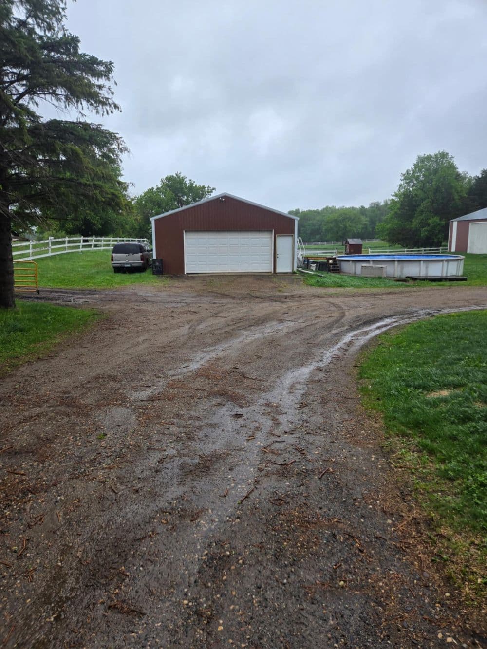 Gravel driveway leading to a brown barn and swimming pool on a rainy day.