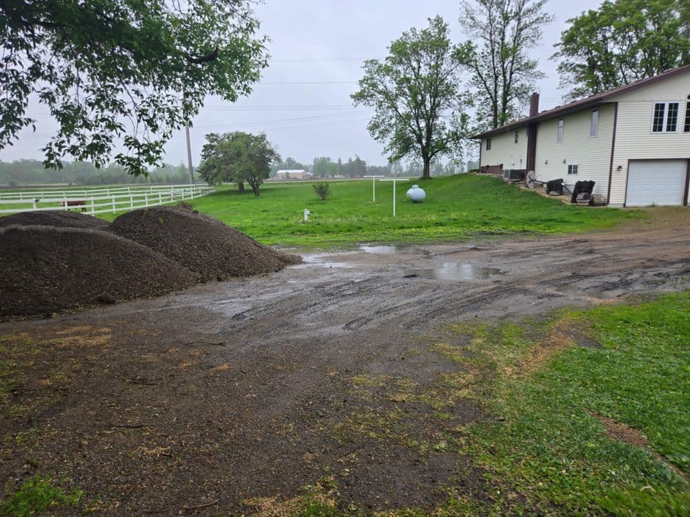 Gravel driveway with wet ground near a house and dirt piles on a cloudy day.