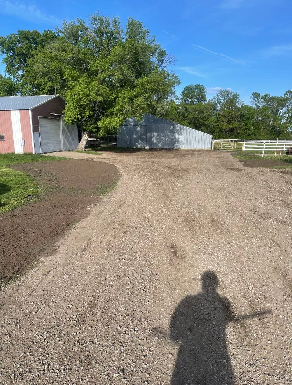 Gravel driveway leading to barns and green trees in a rural setting on a sunny day.