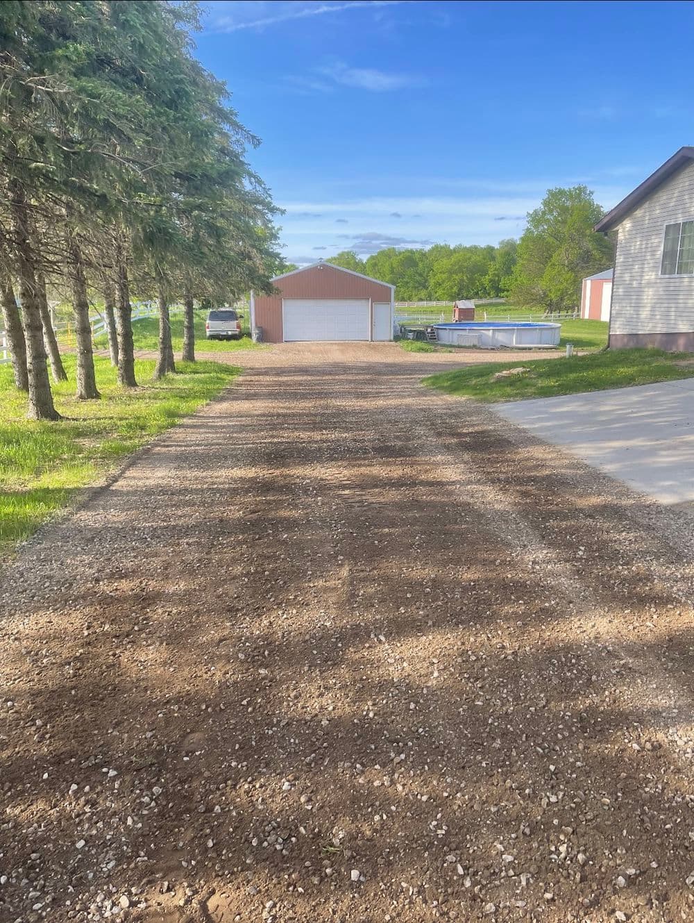 Gravel driveway leading to a shed, surrounded by trees and greenery on a clear day.