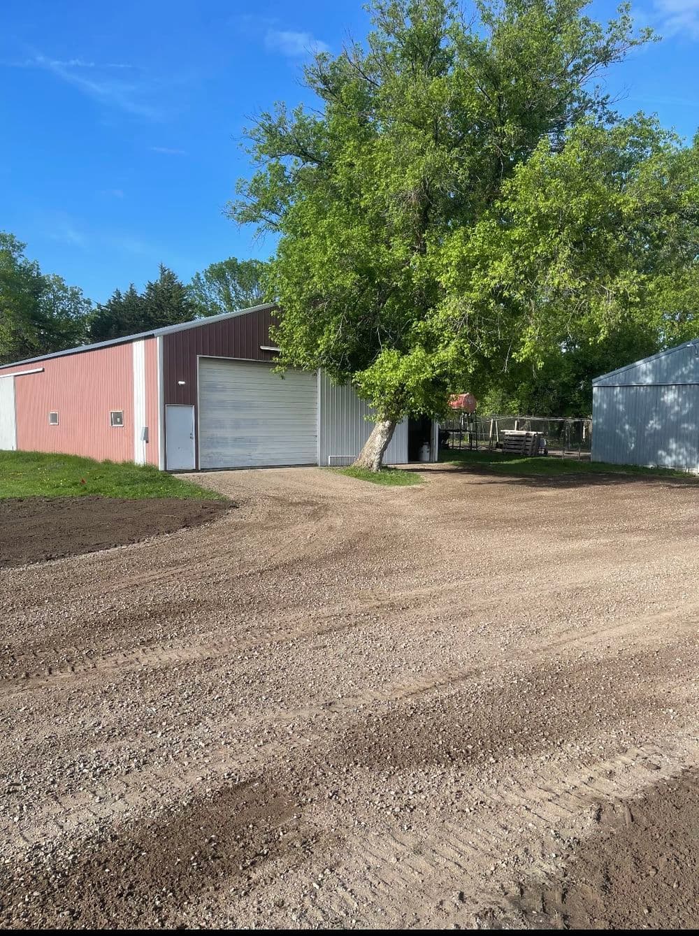 Gravel driveway leading to a red barn and green tree under a blue sky.