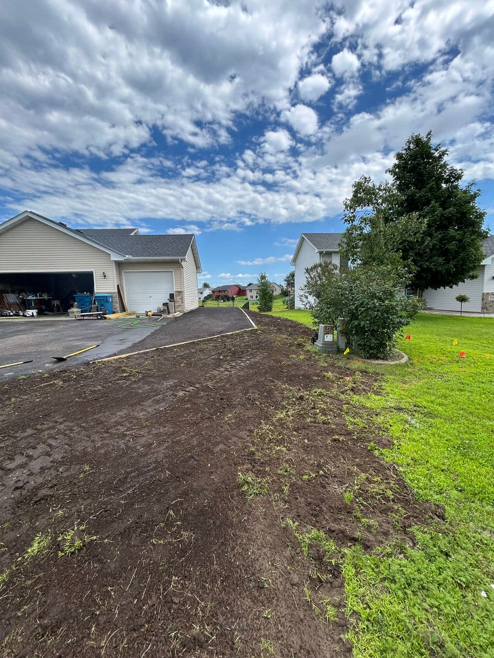 Residential landscaping project with freshly turned earth and clear sky, adjacent homes visible.