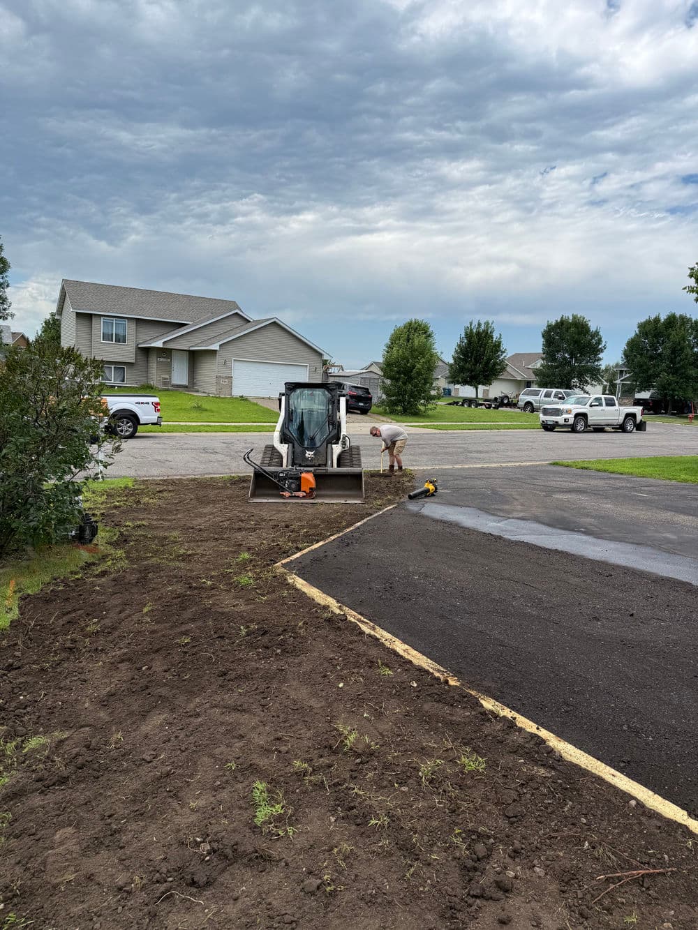 Construction equipment and worker preparing landscape near residential area on a cloudy day.