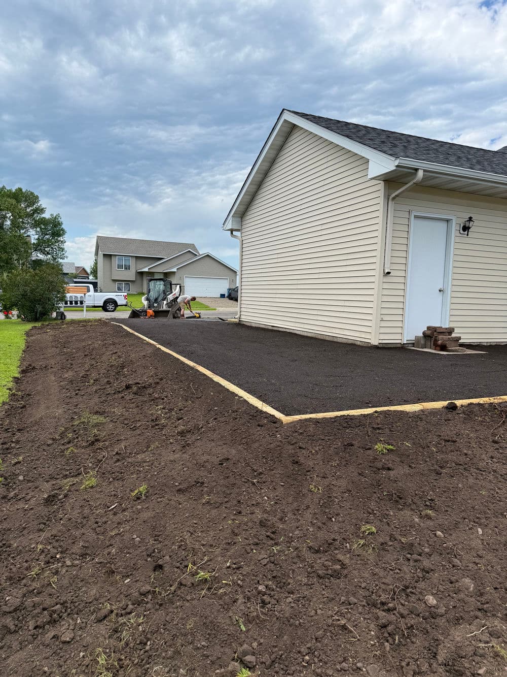Newly laid asphalt driveway beside a house, with freshly graded earth and cloudy sky.