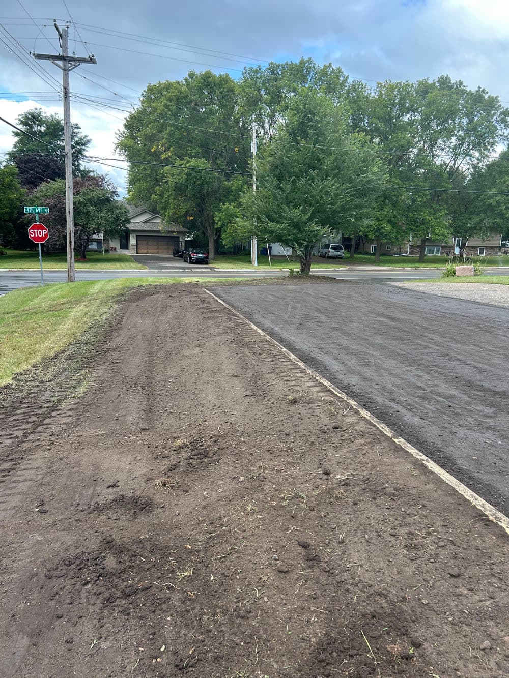 Cleared dirt area next to a grassy sidewalk and stop sign, with trees in the background.
