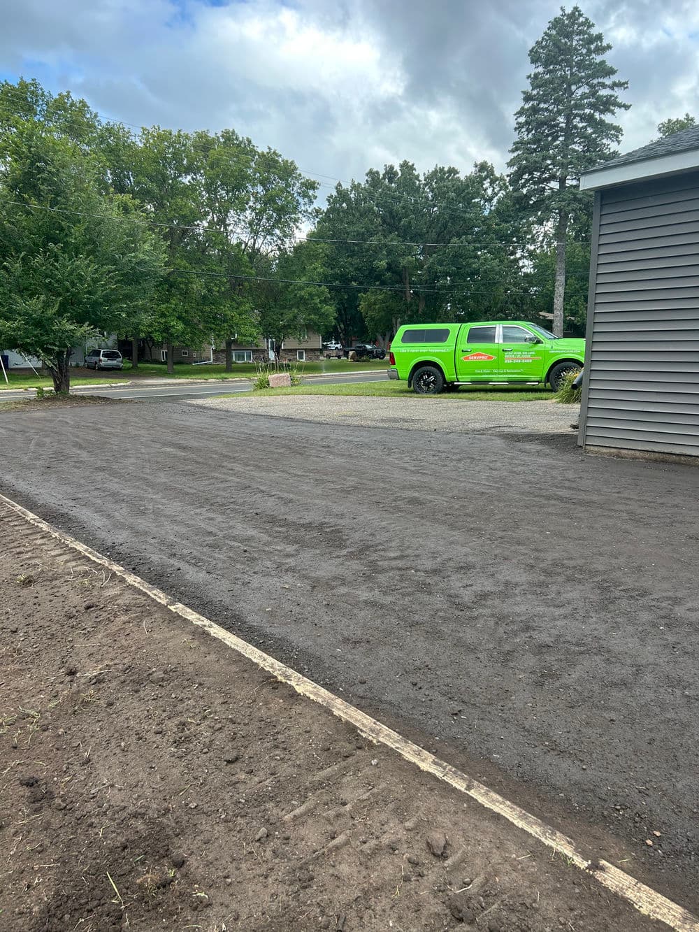 Green vehicle parked near freshly graded driveway in residential area with overcast sky.