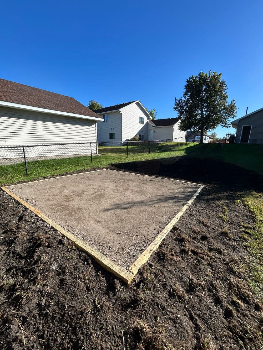 Cleared and leveled backyard site ready for a shed or garden installation. Sunny day, houses nearby.