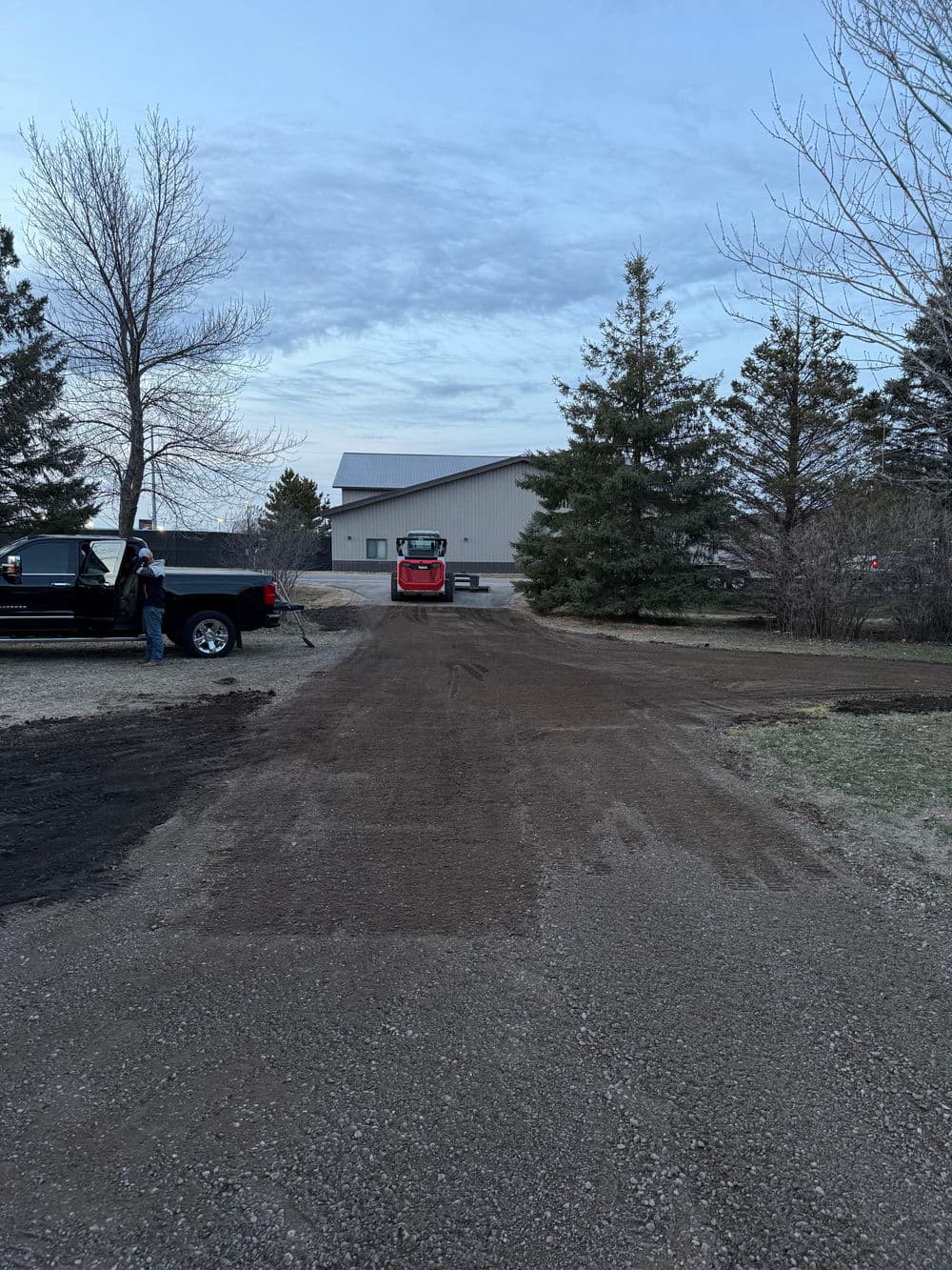 Excavator preparing gravel driveway with a parked truck and house in the background.