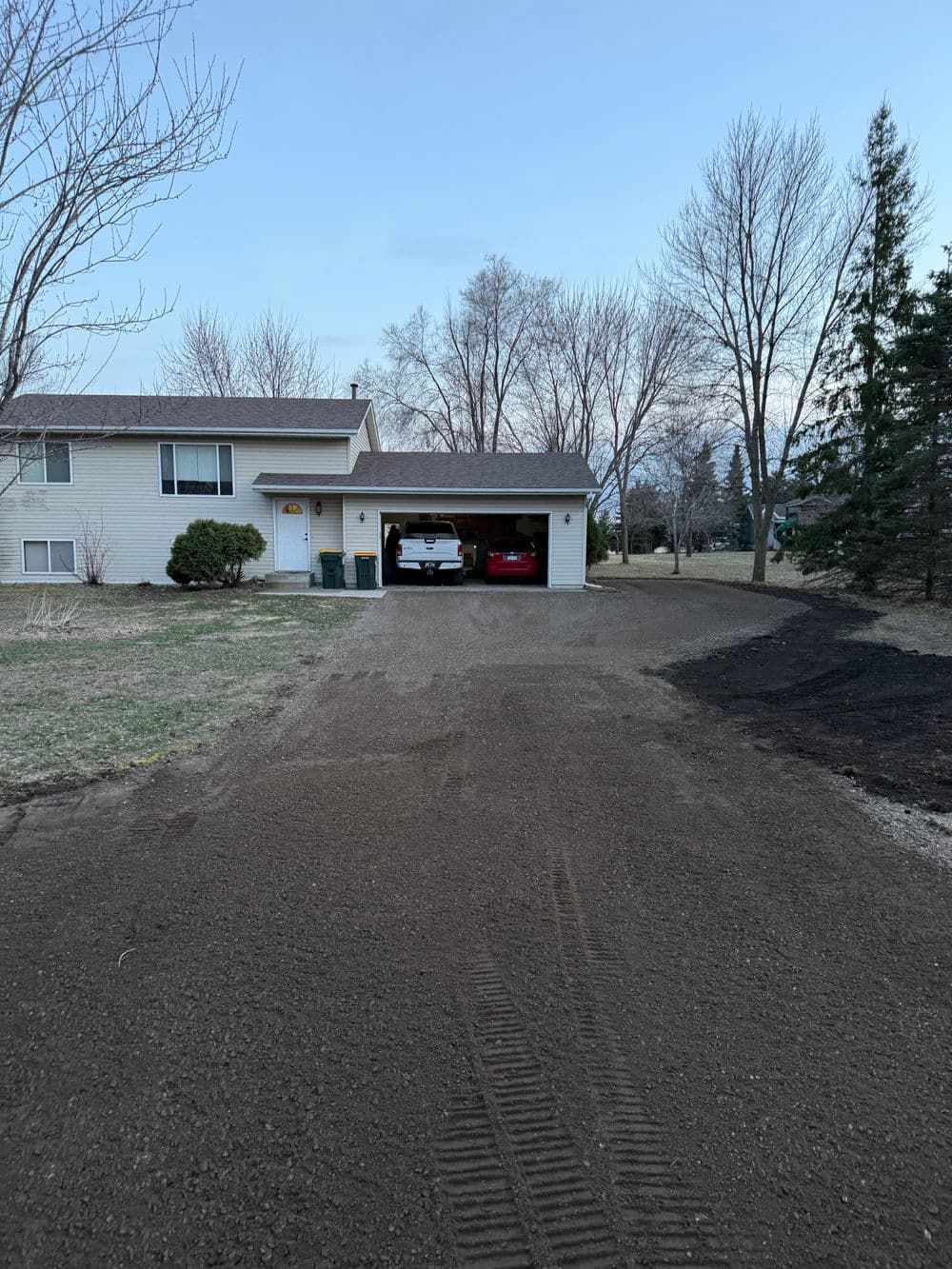 Driveway leading to a house with a garage and parked vehicles surrounded by trees.