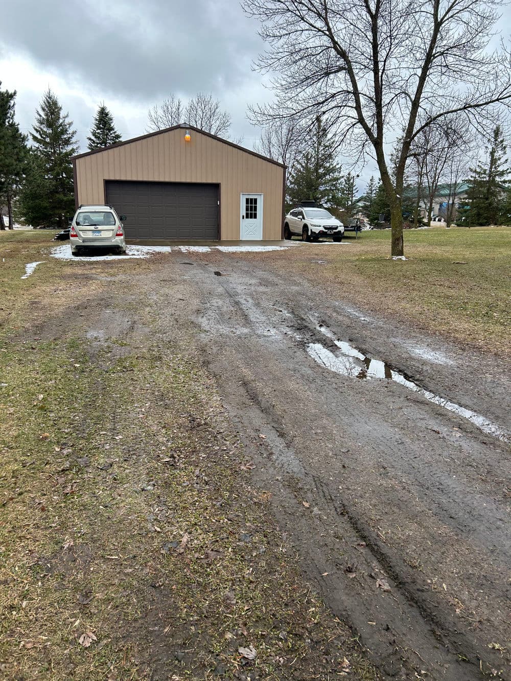 Two cars parked near a muddy driveway and a large metal garage surrounded by trees.