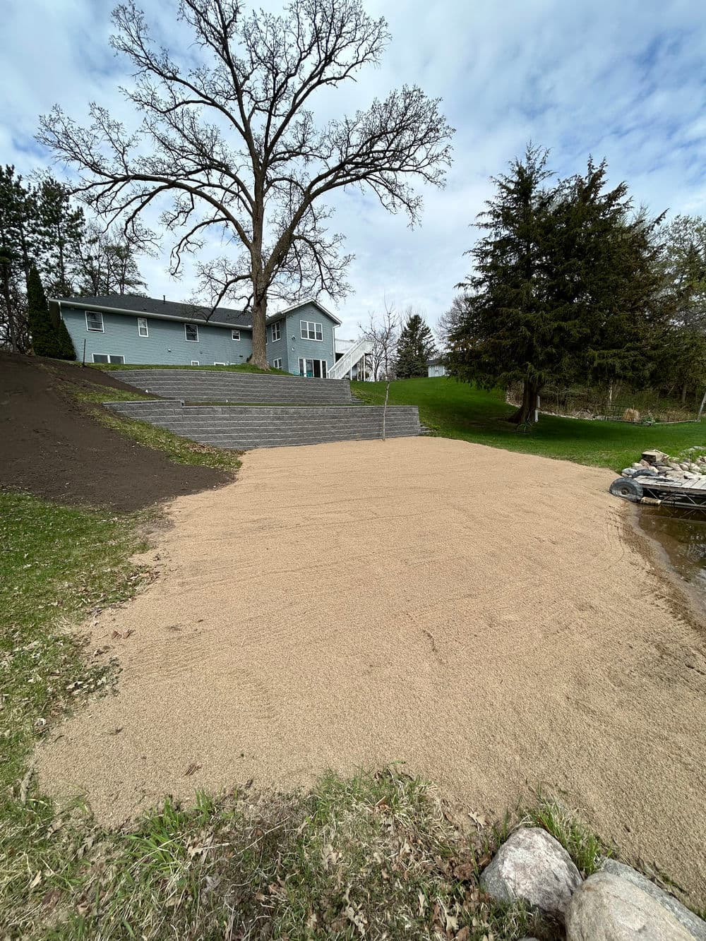 Sandy area with landscaped steps and a green house in the background under a cloudy sky.
