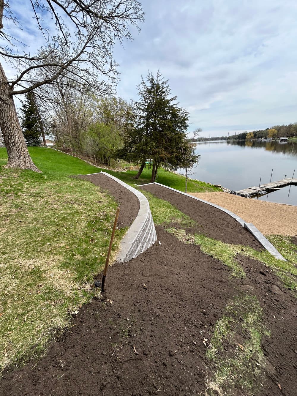 Lakeside landscaping with a curved path, freshly laid soil, and greenery by the water.