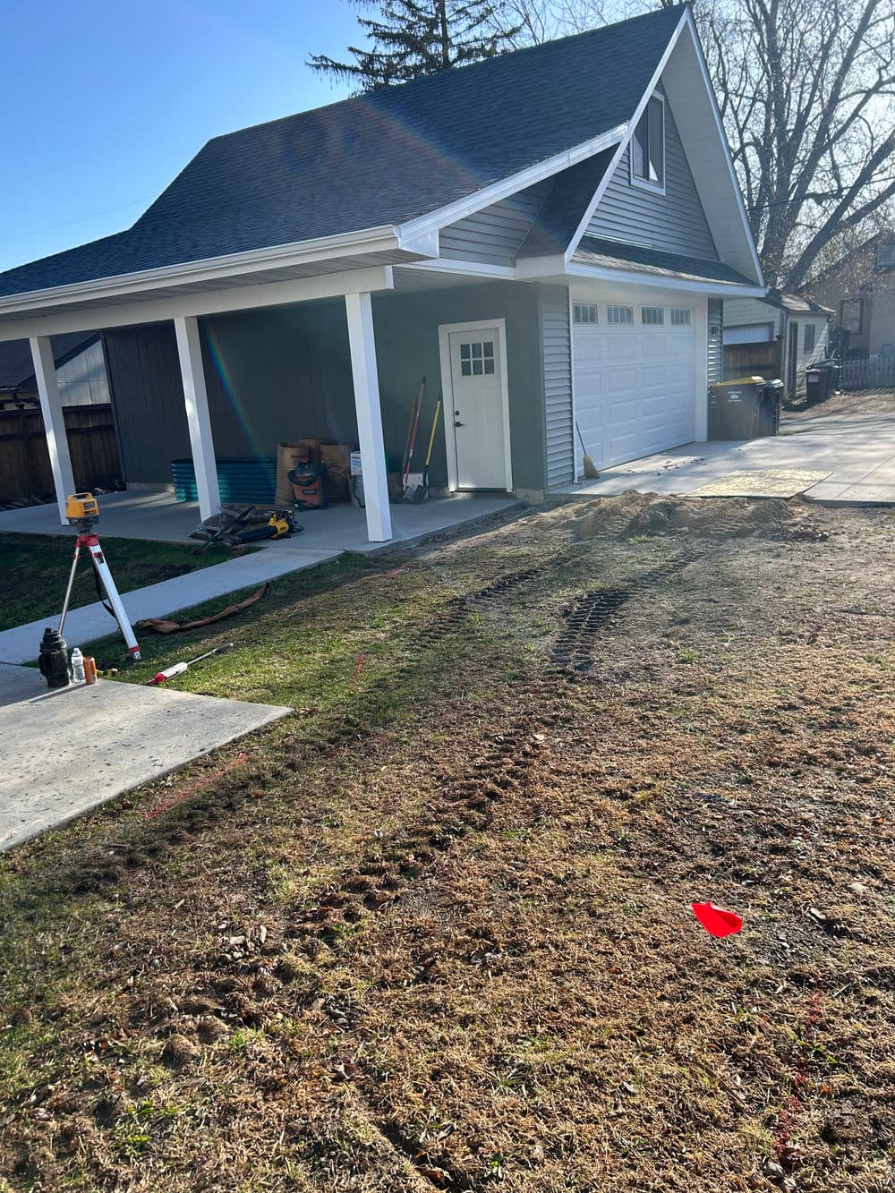 Newly constructed garage with driveway, tools, and tire tracks on grass. Outdoor renovation scene.
