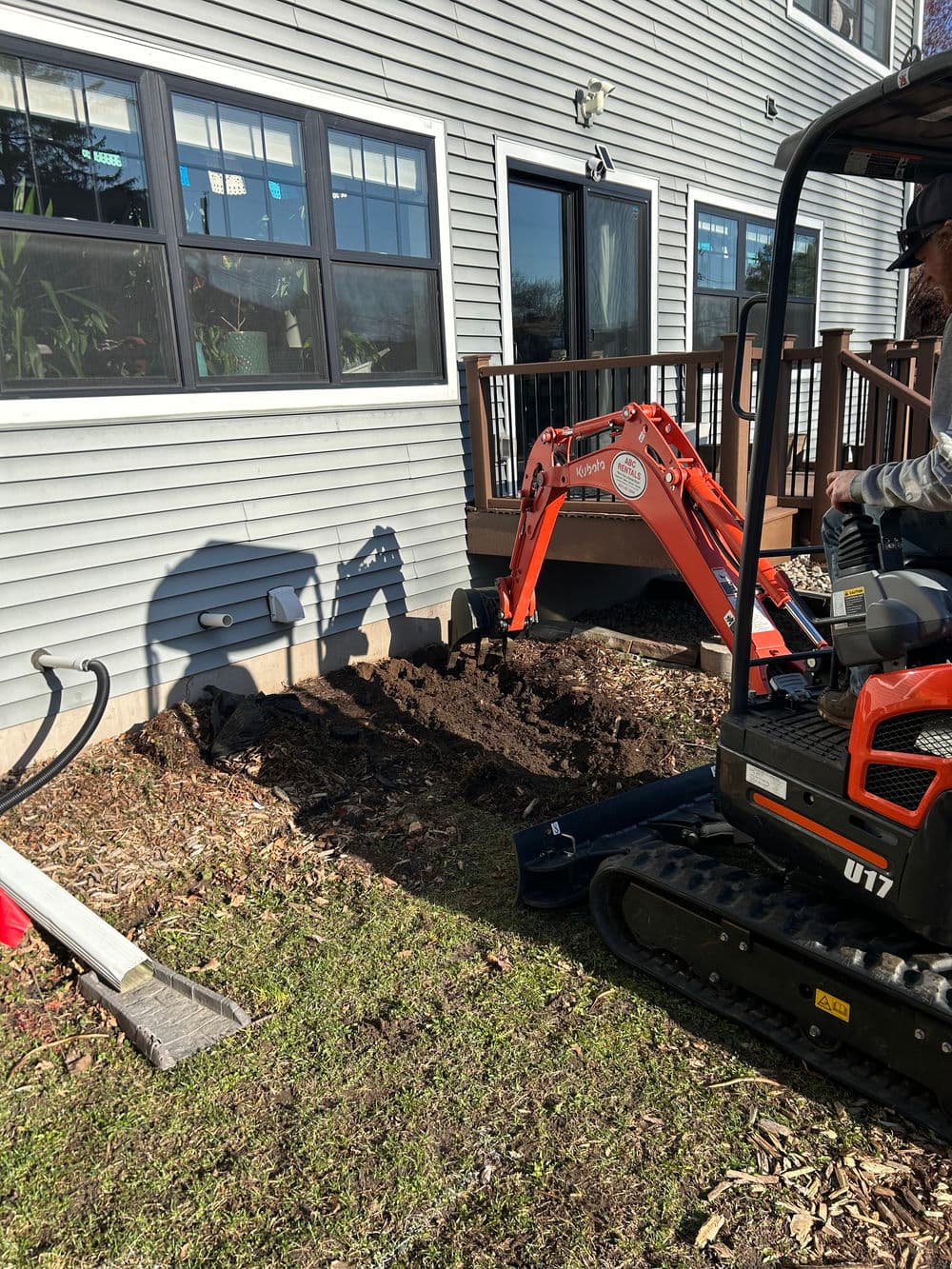 Excavator digging trench near residential home for landscaping or utility work.